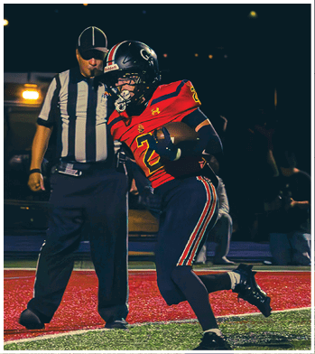 A person wearing a football uniform and helmet is holding a football while another person in a referee uniform stands behind them on a dark field. Description generated by AI