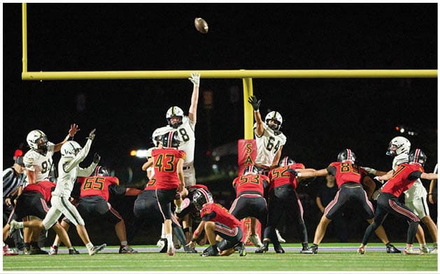 People in football uniforms are on a field with one person jumping to block a kicked football near the goalposts during a nighttime game. Description generated by AI