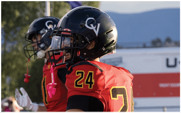 Two people wearing black helmets and red sports jerseys with the number 24 are standing outdoors with a blurred background of trees and mountains. Description generated by AI
