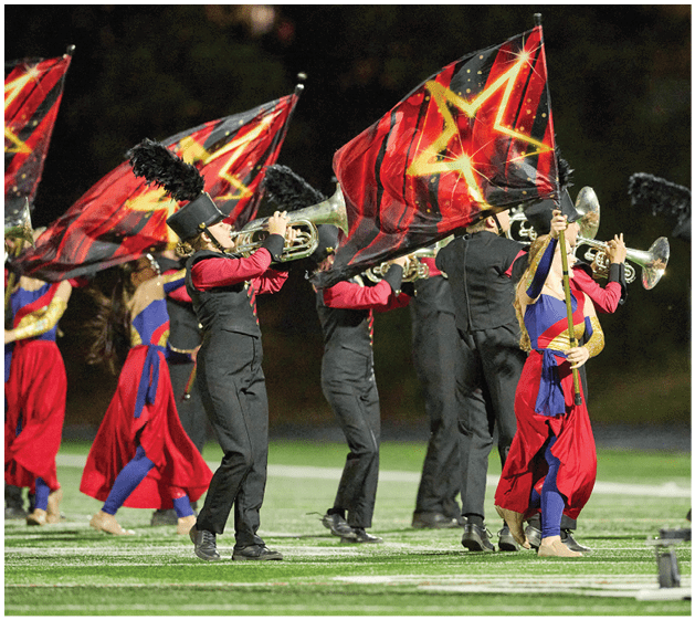 People in marching band uniforms are playing brass instruments and waving red flags with yellow star designs on a football field. Description generated by AI