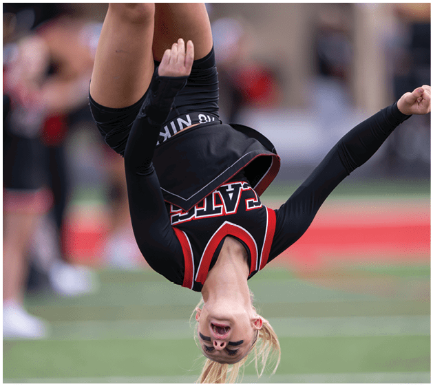 A person wearing a cheerleading uniform is captured mid-flip on a sports field with arms extended. Description generated by AI