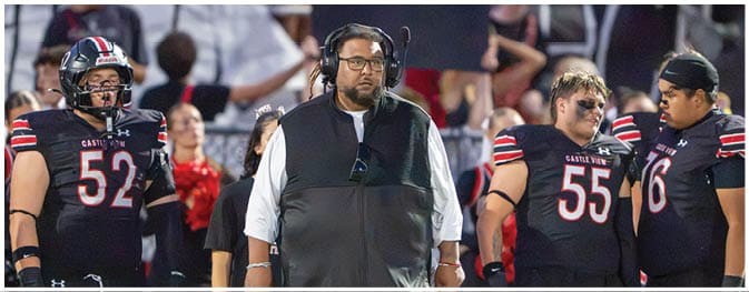 People wearing black football uniforms with red and white stripes stand on a field alongside a person in a black vest and white shirt. Description generated by AI