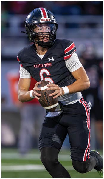 A person wearing a black football uniform with red and white stripes and a helmet is holding a football and appears to be preparing to throw it on a field. Description generated by AI