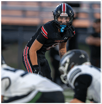 A person in a black football uniform with red and white stripes is crouched and focused on the play while two opposing players in white uniforms are positioned in front of them on a football field. Description generated by AI