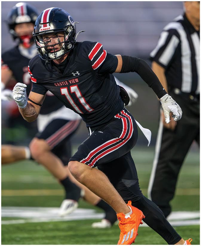 A person in a black football uniform with red and white stripes is running on a field while other players in black and white uniforms are in the background. Description generated by AI