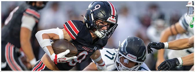 A person wearing a black football uniform with red and white stripes is running with the ball while being pursued by opponents on a football field. Description generated by AI