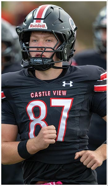 An adult wearing a black football uniform with the number 87 and the words Castle View is standing with hands positioned as if ready to move, with a pink glove tucked into the waistband. Description generated by AI