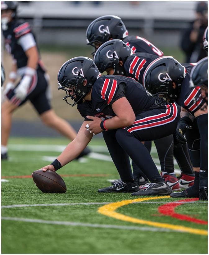 A group of football players in black uniforms with red and white stripes are lined up on a field preparing for a play, with one player holding the football on the ground. Description generated by AI