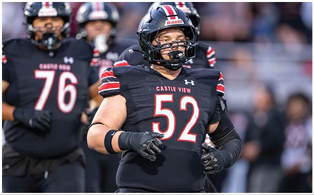 Several people wearing black football uniforms with red and white stripes and helmets are standing on a field, with one person in the foreground wearing number 52 and the words “Castle View" on the jersey. Description generated by AI