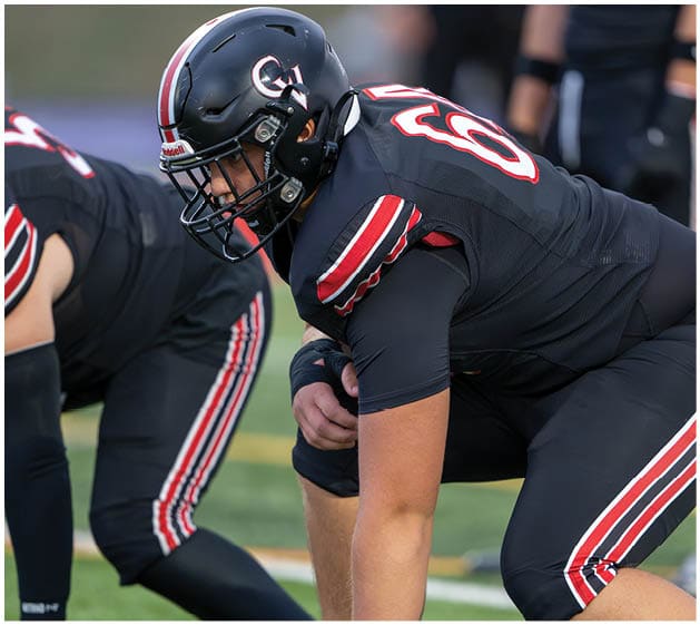 Two people in black football uniforms with red and white stripes are crouched on a green field, preparing for a play. Description generated by AI