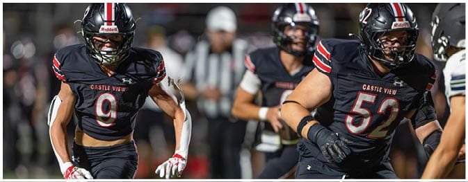 Two people in black football uniforms with red and white stripes and helmets are running on a field during a game. Description generated by AI
