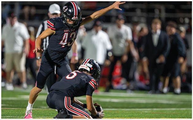 Two people in football uniforms are on a field with one holding the ball on the ground and the other preparing to kick it. Description generated by AI