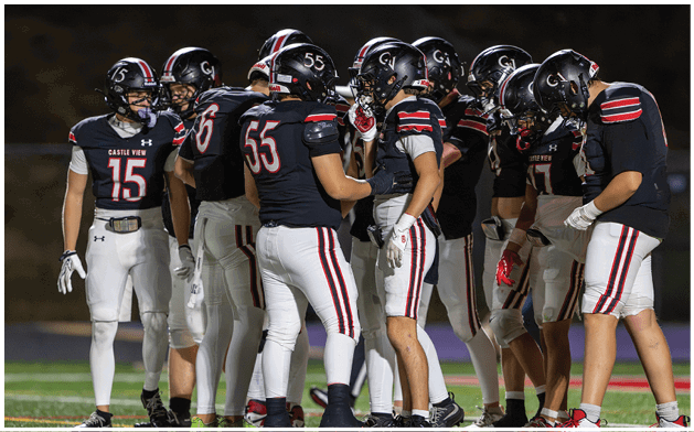 A group of football players in black helmets and dark jerseys with white pants are huddled together on a field during a game. Description generated by AI