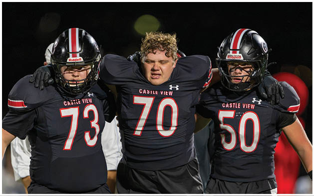 Three people in black football uniforms with helmets stand closely together on a field at night, showing camaraderie. Description generated by AI