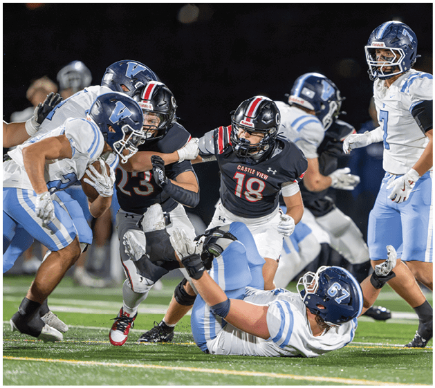 A group of people wearing football uniforms are actively engaged in a play on a field, with one person holding the ball while others attempt to tackle or block. Description generated by AI
