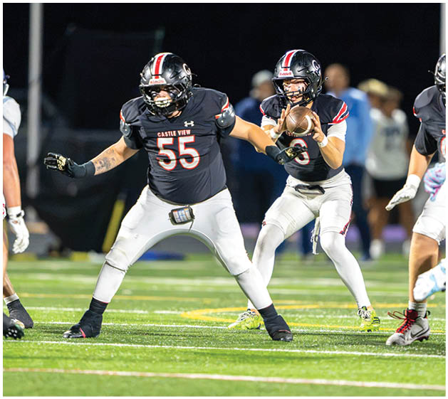 Two people in football uniforms are on a field at night with one holding a football ready to throw and the other positioned in front for protection. Description generated by AI