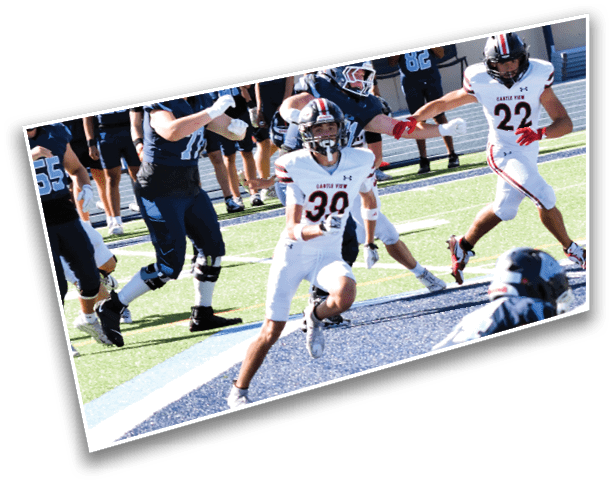 Several people in football uniforms are actively playing on a field with one person running while holding a football. Description generated by AI