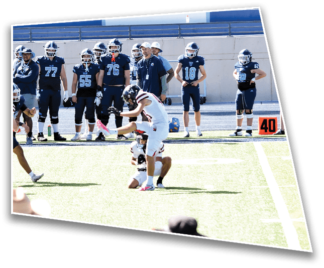 People in football uniforms are playing on a field with others standing on the sidelines and spectators sitting on blue bleachers in the background. Description generated by AI