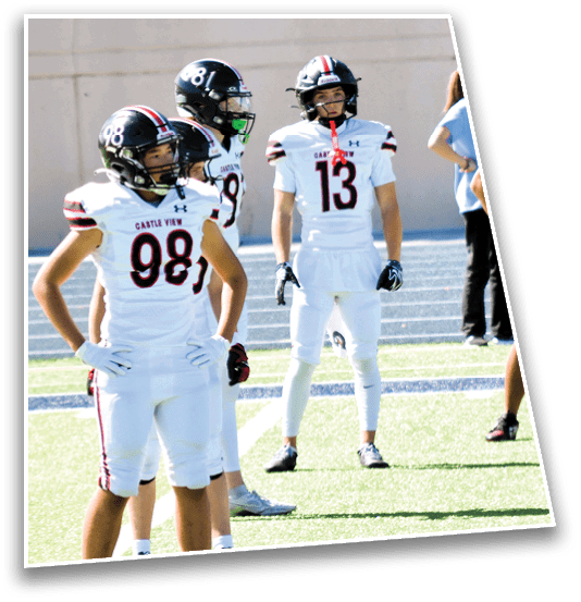 Several people in white football uniforms with helmets stand on a field while a person in casual clothing stands in the background. Description generated by AI