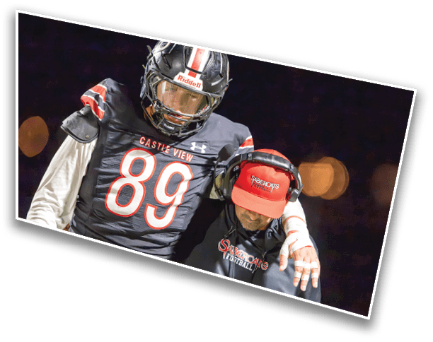 A person wearing a football uniform with the number 89 and a helmet stands next to another person wearing a headset and a red cap with the word “Spartans" on it against a dark background. Description generated by AI