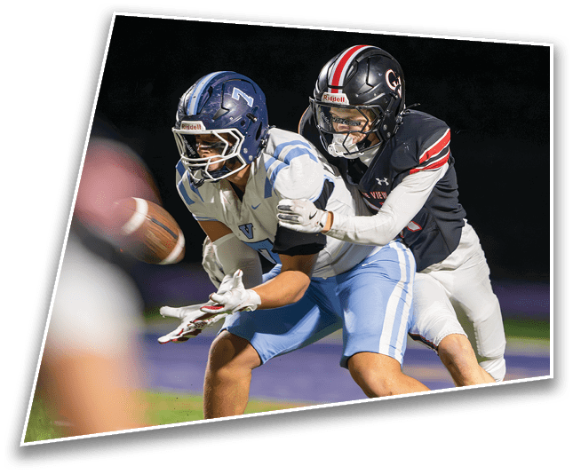 Two people wearing football helmets and uniforms are competing to catch a football in midair on a field at night. Description generated by AI