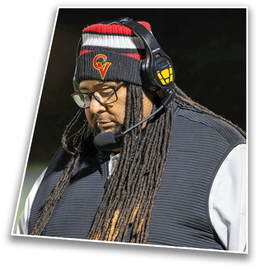 A person with long dreadlocks wearing glasses, a headset, a black and white jacket, and a beanie with a red and yellow logo is standing outdoors, appearing focused. Description generated by AI