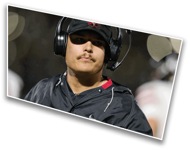 Person wearing a black cap and headset with microphone is looking ahead while dressed in a black jacket, likely coaching or managing a sports event. Description generated by AI