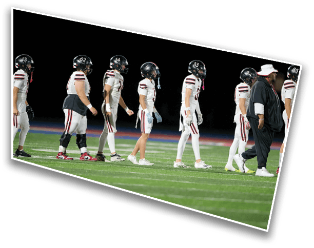 A group of people wearing football uniforms and helmets walk in a line on a field at night alongside a person in a white hat and dark clothing. Description generated by AI