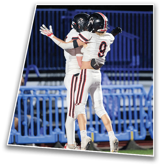 Two people in football uniforms and helmets are celebrating with an embrace on a field with blue bleachers in the background. Description generated by AI