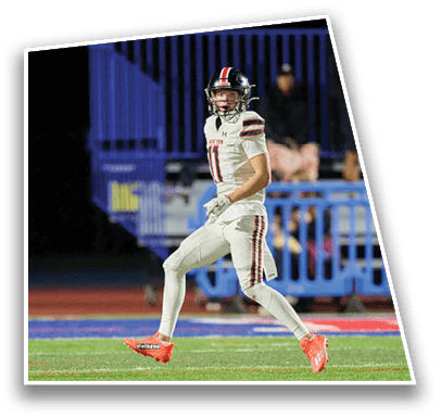 A person wearing a football helmet and white jersey with red and black stripes is looking up at a football in mid-air against a dark background with blue bleachers. Description generated by AI