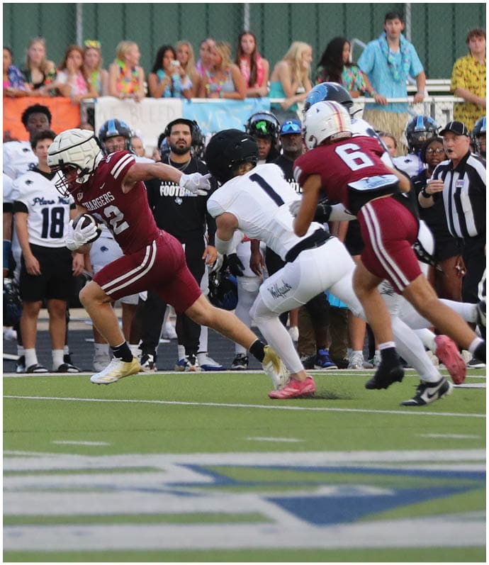 A group of people in football uniforms are actively playing on a field with spectators and a team standing on the sidelines in the background. Description generated by AI