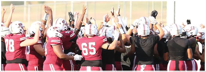 A group of people wearing maroon and white sports uniforms are gathered closely together on a field with their hands raised in a celebratory or motivational gesture. Description generated by AI