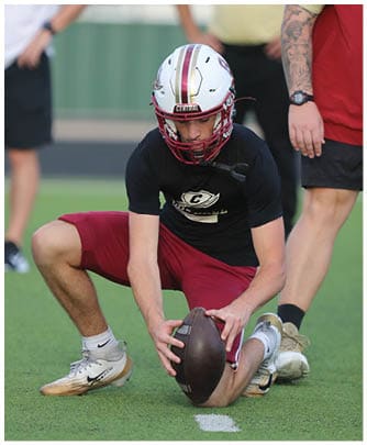 A person wearing a white football helmet with red and gold stripes and the word “CENTRAL" on the front is standing with hands on hips against a blurred background. Description generated by AI