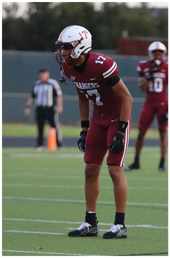 A person wearing a football helmet and maroon jersey with the number 17 is on a field with trees and a fence in the background. Description generated by AI