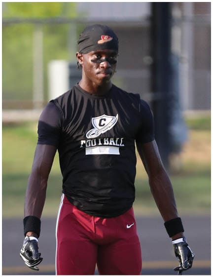 A person wearing a black cap, black shirt with a football logo, red shorts, and gloves stands on a sports field with a scoreboard in the background. Description generated by AI