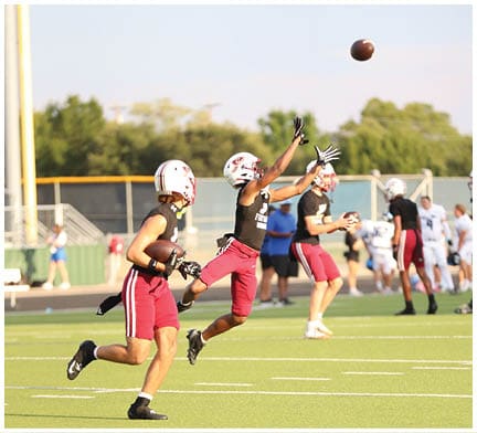 Several people wearing helmets and sports uniforms are playing football outdoors with one person reaching up to catch a football in midair. Description generated by AI