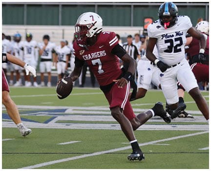 A person in a maroon football uniform is running with the ball while being chased by several people in white uniforms on a football field with spectators in the background. Description generated by AI