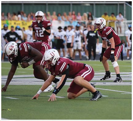 Two people in maroon sports uniforms are crouched on a football field in a ready position facing an opponent in a white uniform. Description generated by AI