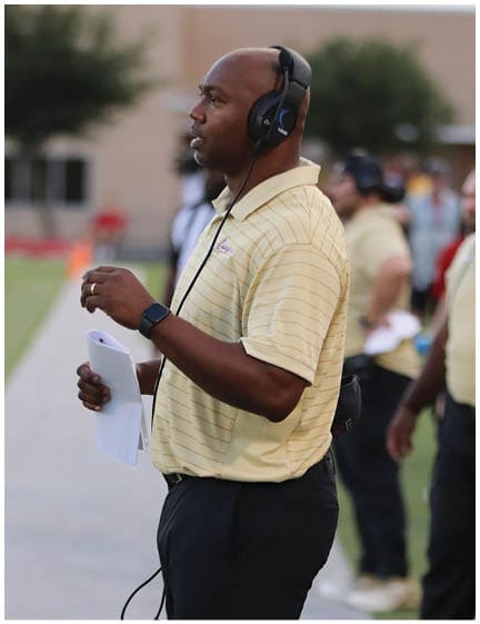 A person wearing a yellow striped polo shirt and black pants is holding papers and wearing a headset while standing outdoors. Description generated by AI
