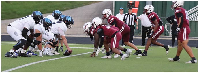 Two opposing American football teams in helmets and uniforms are lined up facing each other on a field with referees and spectators in the background. Description generated by AI
