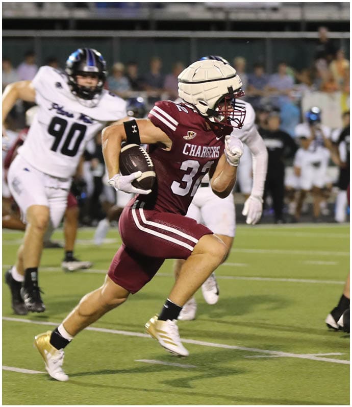 People wearing maroon and white football uniforms are actively playing on a green field with blurred spectators in the background. Description generated by AI