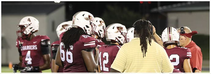 Several people wearing football uniforms and helmets are gathered on a field with one person in a yellow striped shirt interacting with them. Description generated by AI