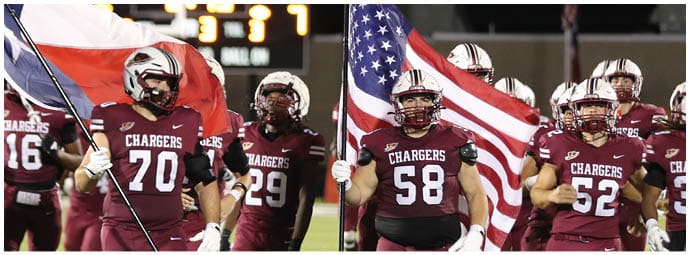 People in football uniforms holding flags stand on a field with a scoreboard visible in the background. Description generated by AI