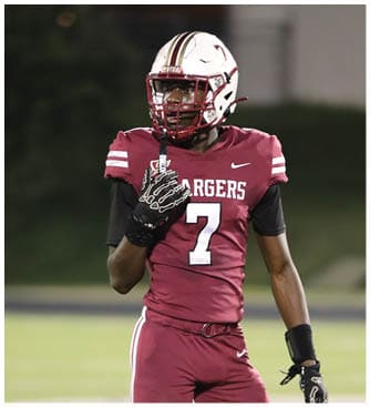 A person wearing a maroon sports jersey and a white football helmet with maroon stripes is on a field at night. Description generated by AI