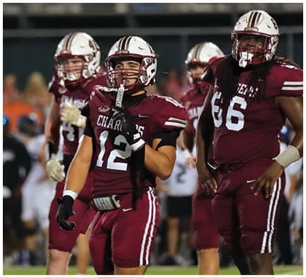 Two people in football uniforms stand on a field with one wearing a white jersey numbered 72 and the other in a maroon jersey, with blurred figures and spectators in the background. Description generated by AI