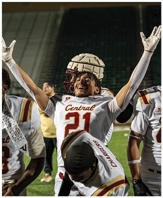 A group of football players in white uniforms celebrate on a field with one player raising their arms joyfully. Description generated by AI