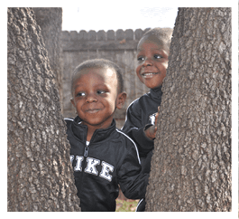 Two children wearing black Nike jackets are smiling and peeking out from behind tree trunks in an outdoor setting. Description generated by AI