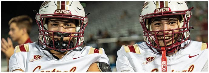 Two people wearing white football jerseys with “Central" written on them and red helmets are standing side by side. Description generated by AI