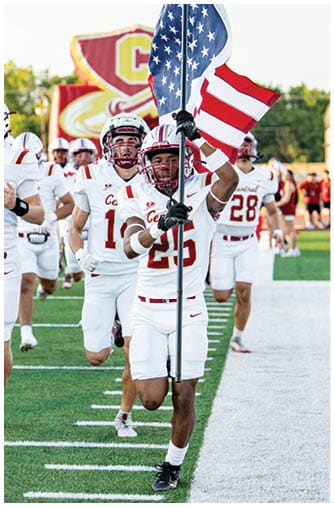 A group of people wearing white sports uniforms with red lettering are running on a green football field. Description generated by AI