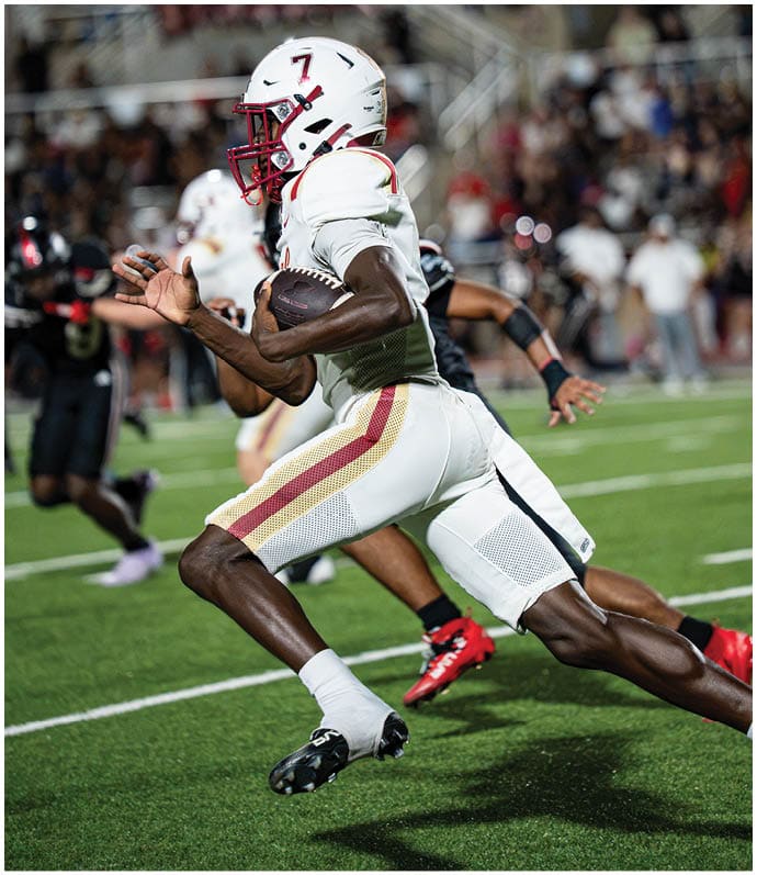 An athlete wearing a white football uniform with red and gold stripes is running on a green field while holding a football, with other players and a crowd visible in the background. Description generated by AI